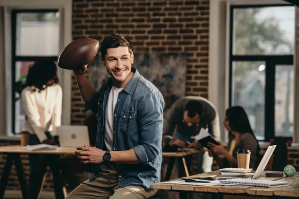 Glücklicher junger Mann mit Rugbyball im modernen Loft-Büro mit Kollegen, die im Hintergrund arbeiten — Stockfoto