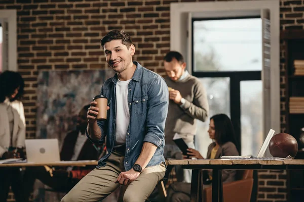 Улыбающийся молодой человек, держащий чашку кофе и смотрящий на камеру в современном лофт-офисе с коллегами на заднем плане — стоковое фото