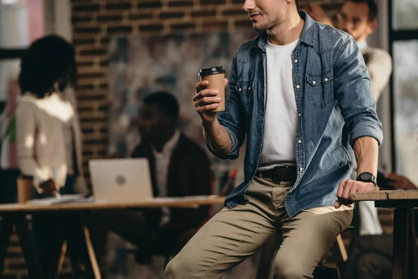 Cropped view of young man holding cup of coffee in modern loft office with colleagues working on background — Stock Photo