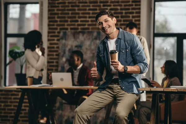 Молодой человек держит кофе и дает большие пальцы вверх вывеску в современном офисе лофт с коллегами на заднем плане — стоковое фото