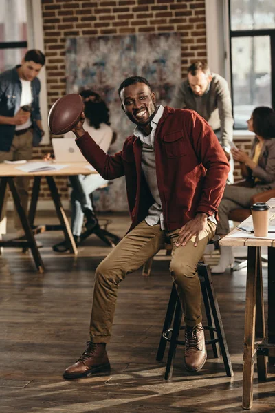 Sorridente uomo d'affari africano americano casual che tiene la palla da rugby con i colleghi che lavorano dietro in ufficio loft — Foto stock