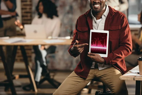 Recortado vista de sonriente afroamericano casual hombre de negocios celebración de la tableta con gráfico en la pantalla y colegas que trabajan detrás en la oficina loft - foto de stock