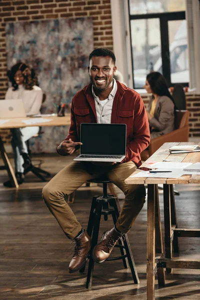 Hombre de negocios casual afroamericano sosteniendo portátil con pantalla en blanco y colegas que trabajan detrás en la oficina loft - foto de stock