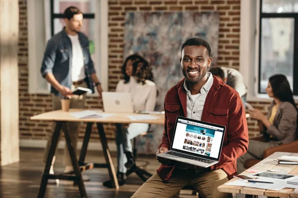 African american casual businessman holding laptop with shopping web site and colleagues working behind in loft office — Stock Photo
