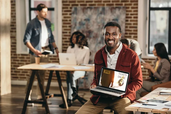 Africano americano casual hombre de negocios celebración de ordenador portátil con entradas en línea sitio web y colegas que trabajan detrás en la oficina loft - foto de stock