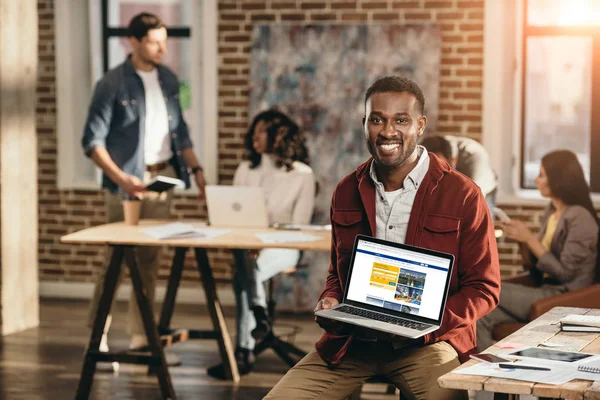 African american casual businessman holding laptop with booking web site and colleagues working behind in loft office — Stock Photo