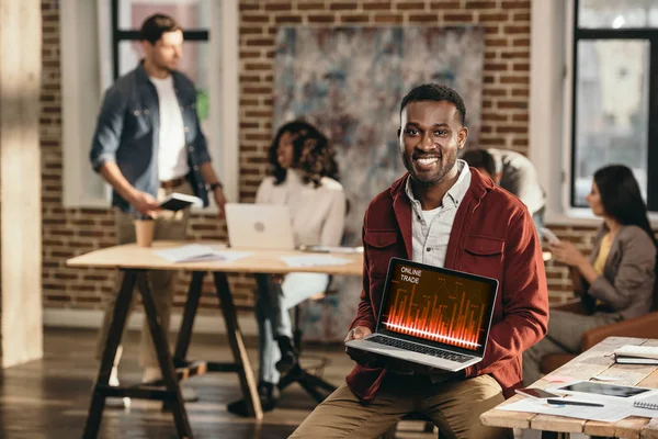 African american casual businessman holding laptop with graph on screen and colleagues working behind in loft office — Stock Photo