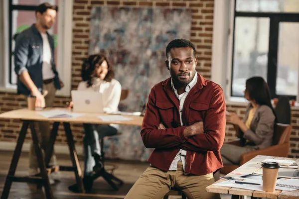 Афро-американський випадковий бізнесмен, що сидить зі схрещеними руками, і колеги працюють позаду в офісі лофт — стокове фото