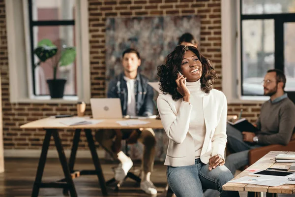 Весела афроамериканська випадкова бізнес-леді сидить і розмовляє на смартфоні в офісі лофт з колегами, які працюють позаду — стокове фото
