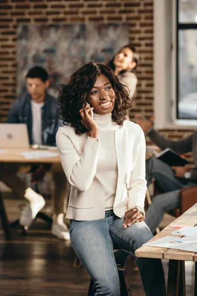 Cheerful african american casual businesswoman sitting and talking on smartphone in loft office with colleagues working behind — Stock Photo