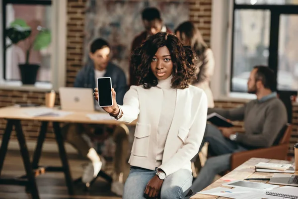 Grave afro-américaine casual femme d'affaires pointant vers smartphone avec écran blanc dans le bureau loft avec des collègues derrière — Photo de stock
