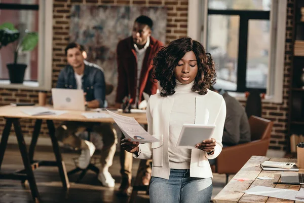 Mujer de negocios casual afroamericana seria sosteniendo papeles con colegas que trabajan detrás en la oficina del desván - foto de stock