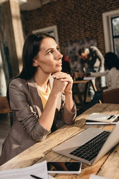 Mujer de negocios pensativa sentada en el escritorio con el ordenador portátil y trabajando en la oficina loft con colegas de fondo - foto de stock
