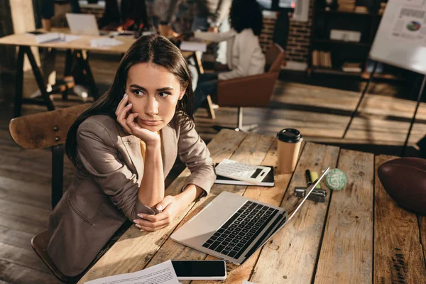 Nachdenkliche Geschäftsfrau sitzt am Schreibtisch mit Laptop und arbeitet im Loftbüro an einem Projekt mit Kollegen im Hintergrund — Stockfoto