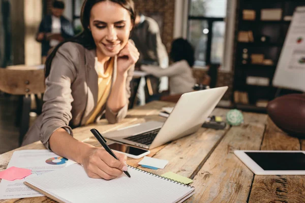 Bella donna d'affari di successo seduta alla scrivania con computer portatile e che lavora su progetto in ufficio loft con colleghi sullo sfondo — Foto stock