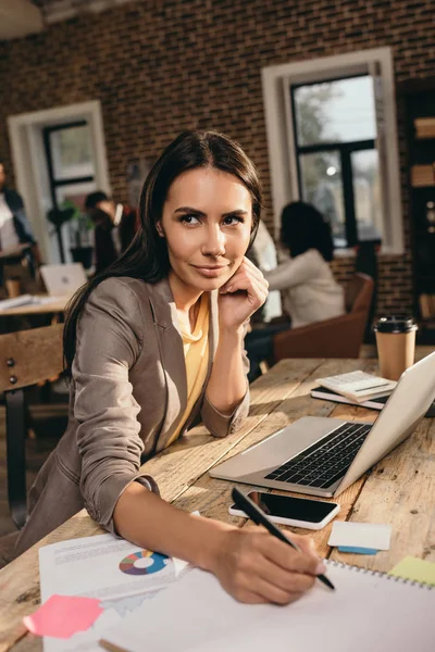 Donna d'affari di successo seduta alla scrivania con computer portatile e che lavora a progetto a ufficio loft con colleghi su sfondo — Foto stock