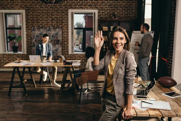 Smiling business woman waving with hand at loft office with colleagues on background — Stock Photo