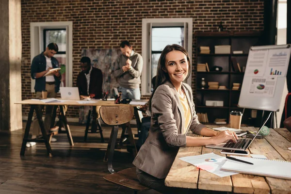 Femme d'affaires souriante assise au bureau avec ordinateur portable et travaillant sur un projet au bureau loft avec des collègues sur fond — Photo de stock