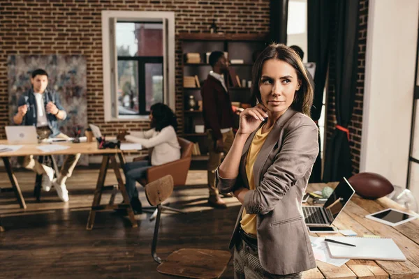 Pensive business woman standing at desk with laptop at loft office with colleagues on background — Stock Photo