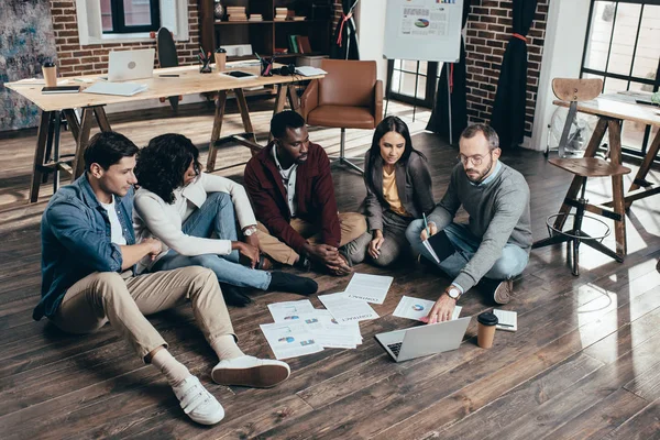 Grupo multiétnico centrado de compañeros de trabajo sentados en el piso y discutir juntos nuevo proyecto en la oficina loft moderna - foto de stock