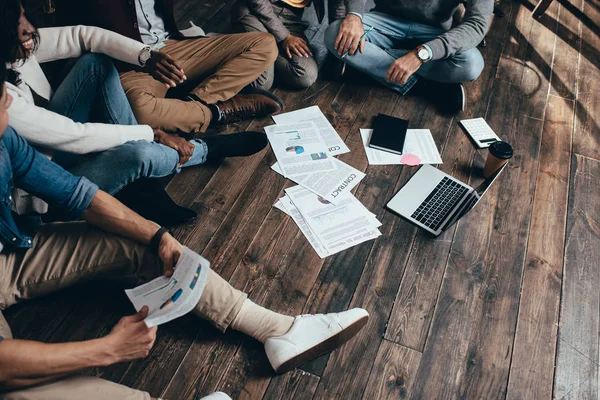 Abgeschnittene Ansicht einer multiethnischen Gruppe von Kollegen, die auf dem Boden sitzen und gemeinsam an einem neuen Projekt arbeiten — Stockfoto