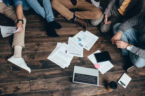 Abgeschnittene Ansicht einer multiethnischen Gruppe von Kollegen, die auf dem Boden sitzen und gemeinsam an einem neuen Projekt arbeiten — Stockfoto