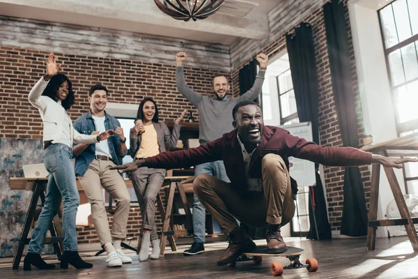 Group of happy multiethnic coworkers having fun with skateboard in loft office — Stock Photo