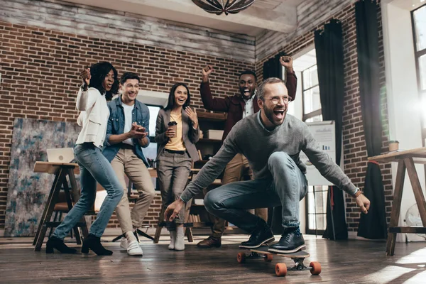 Grupo de compañeros de trabajo multiétnicos divirtiéndose con monopatín en la oficina loft - foto de stock