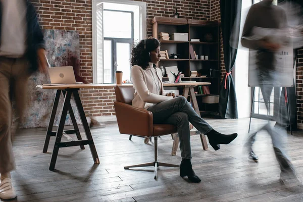 Femme d'affaires afro-américaine dans des heaphones assis sur la chaise avec des collègues en mouvement flou dans le bureau loft moderne — Photo de stock