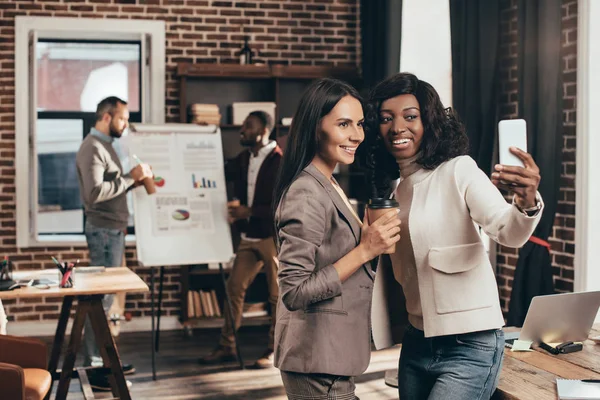 Pareja multiétnica de mujeres de negocios tomando selfie en el teléfono inteligente en la oficina loft con colegas de fondo - foto de stock
