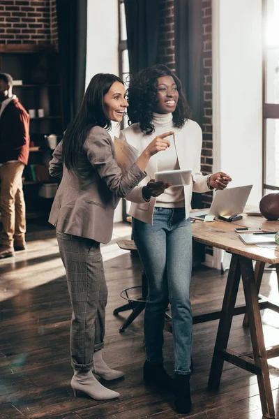 Coppia multietnica di donne d'affari che sorridono e lavorano insieme in ufficio loft con colleghi sullo sfondo — Foto stock