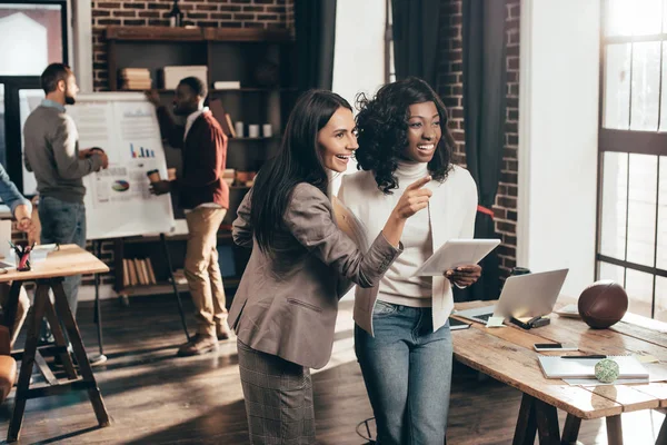 Coppia multietnica di donne d'affari che sorridono e lavorano in ufficio loft con colleghi sullo sfondo — Foto stock