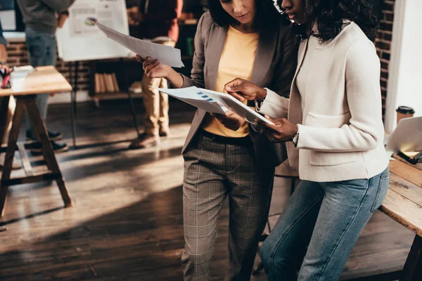 Cropped view of multiethnic couple of business women working in loft office with colleagues on background — Stock Photo