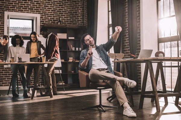 Homem de negócios casual tomando selfie no smartphone com grupo multiétnico de colegas de trabalho atrás no escritório loft moderno — Fotografia de Stock