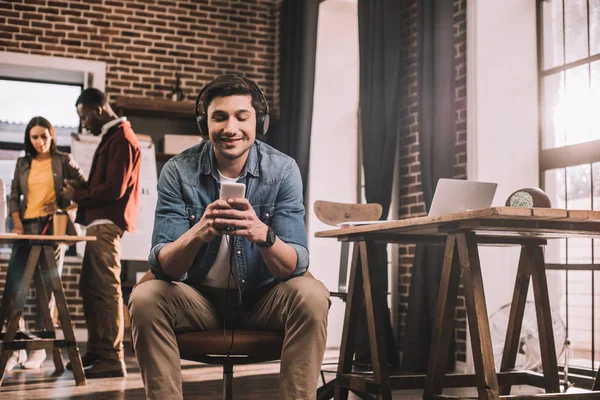 Случайный улыбающийся бизнесмен с помощью смартфона с мультиэтнической группой рабочих коллег позади в современном лофт-офисе — стоковое фото