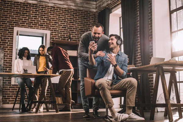 Deux hommes souriants utilisant un smartphone avec un groupe multiethnique d'hommes d'affaires occasionnels dans le bureau loft moderne derrière — Photo de stock