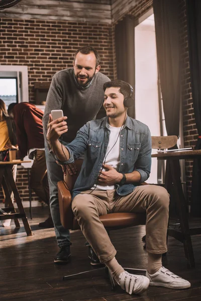 Dos hombres sonrientes usando teléfono inteligente con colegas que trabajan detrás en la oficina moderna loft - foto de stock