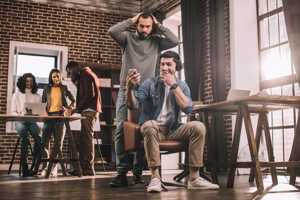 Dos hombres sorprendidos usando teléfono inteligente con grupo multiétnico de empresarios casuales que trabajan en la oficina loft moderna detrás - foto de stock