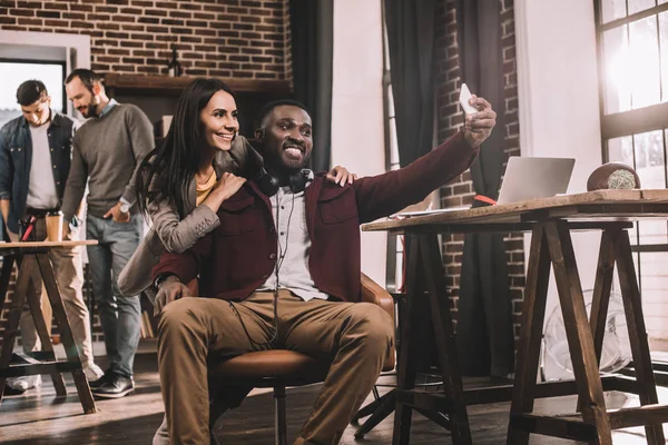 Couple de collègues prenant selfie en utilisant un smartphone avec deux collègues en arrière-plan — Photo de stock