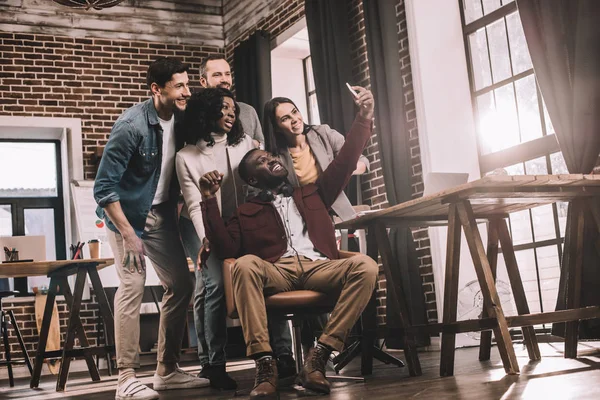 Grupo de alegres compañeros de trabajo tomando selfie usando teléfono inteligente en la oficina loft con retroiluminación - foto de stock