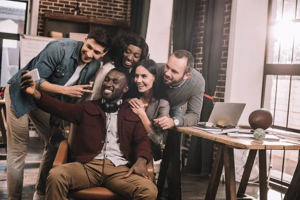 Groupe de collègues multiethniques joyeux prenant selfie en utilisant un smartphone dans le bureau loft avec rétro-éclairé — Photo de stock