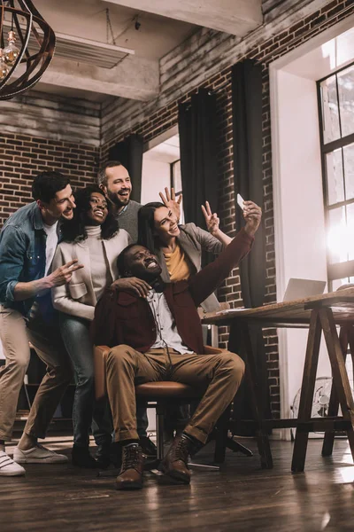 Gruppo di allegri colleghi multietnici scattare selfie utilizzando smartphone in ufficio soppalco con retroilluminazione — Foto stock