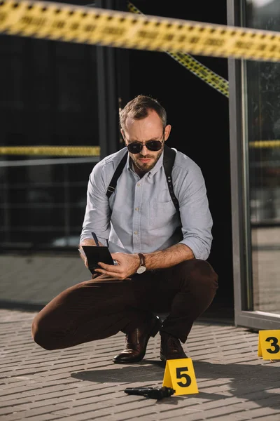 Sitzender männlicher Kriminalbeamter schaut auf die Waffe und macht sich Notizen am Tatort — Stockfoto