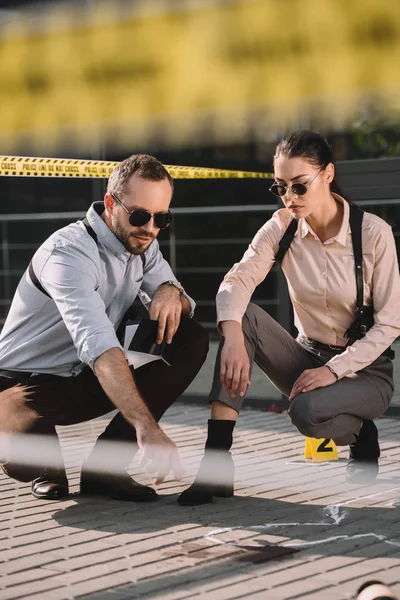 Male and female detectives sitting and looking at chalk line at crime scene — Stock Photo