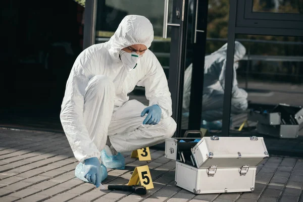 Criminólogo masculino con traje protector y guantes de látex sentado cerca de la pistola de pruebas - foto de stock