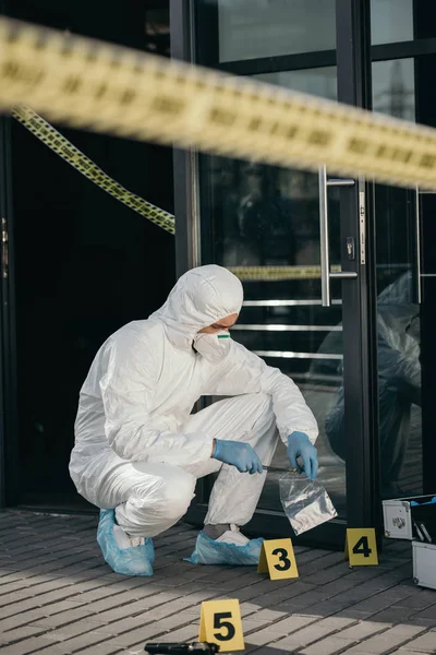 Criminologista do sexo masculino em terno de proteção e luvas de látex embalando provas com wizzles na cena do crime — Fotografia de Stock