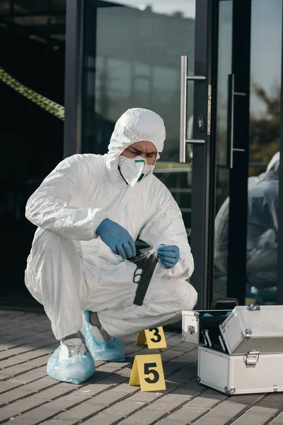 Male criminologist in protective suit and latex gloves sitting with evidence gun in arms — Stock Photo