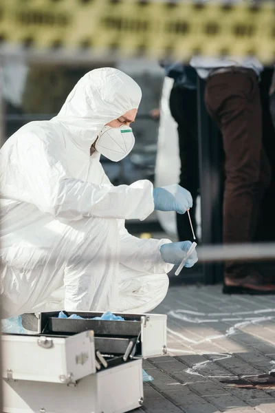 Male criminologist in protective suit and latex gloves collecting blood sample in test tube at crime scene — Stock Photo