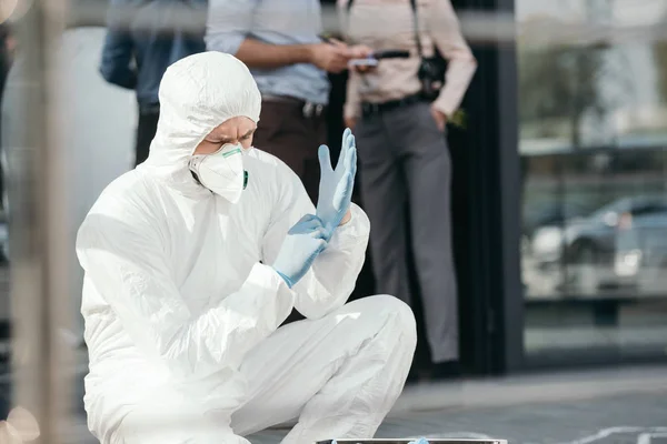 Male criminologist in protective wearing latex gloves with cropped detectives behind — Stock Photo