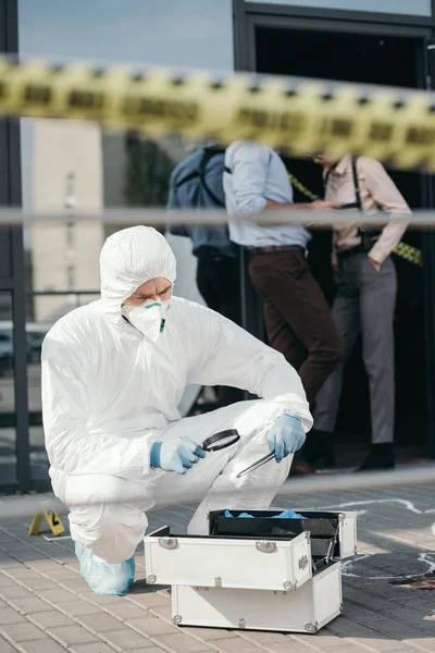 Criminologue masculin en combinaison de protection et gants en latex regardant à travers une loupe à des preuves importantes — Photo de stock